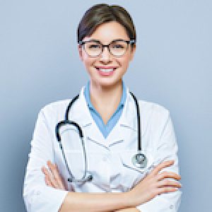 Portrait of glad smiling doctor in white uniform standing with crossed hands on blue clinic background